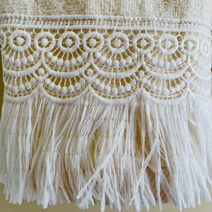 May include: A white towel with a white crocheted lace trim and fringe. The fringe is long and flowing.