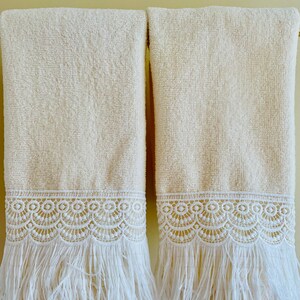 May include: Two cream-colored towels with white lace trim and fringe. The towels are hanging on a wall.