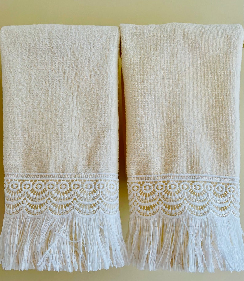 May include: Two cream-colored towels with white lace trim and fringe. The towels are hanging on a wall.