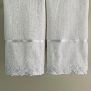 May include: Two white bath towels with a white satin ribbon and white embroidered lace trim.