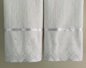 May include: Two white bath towels with a white satin ribbon and white embroidered lace trim.