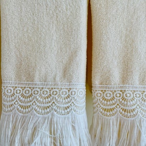 May include: Two cream-colored towels with white lace trim and fringe. The towels are hanging on a wall.