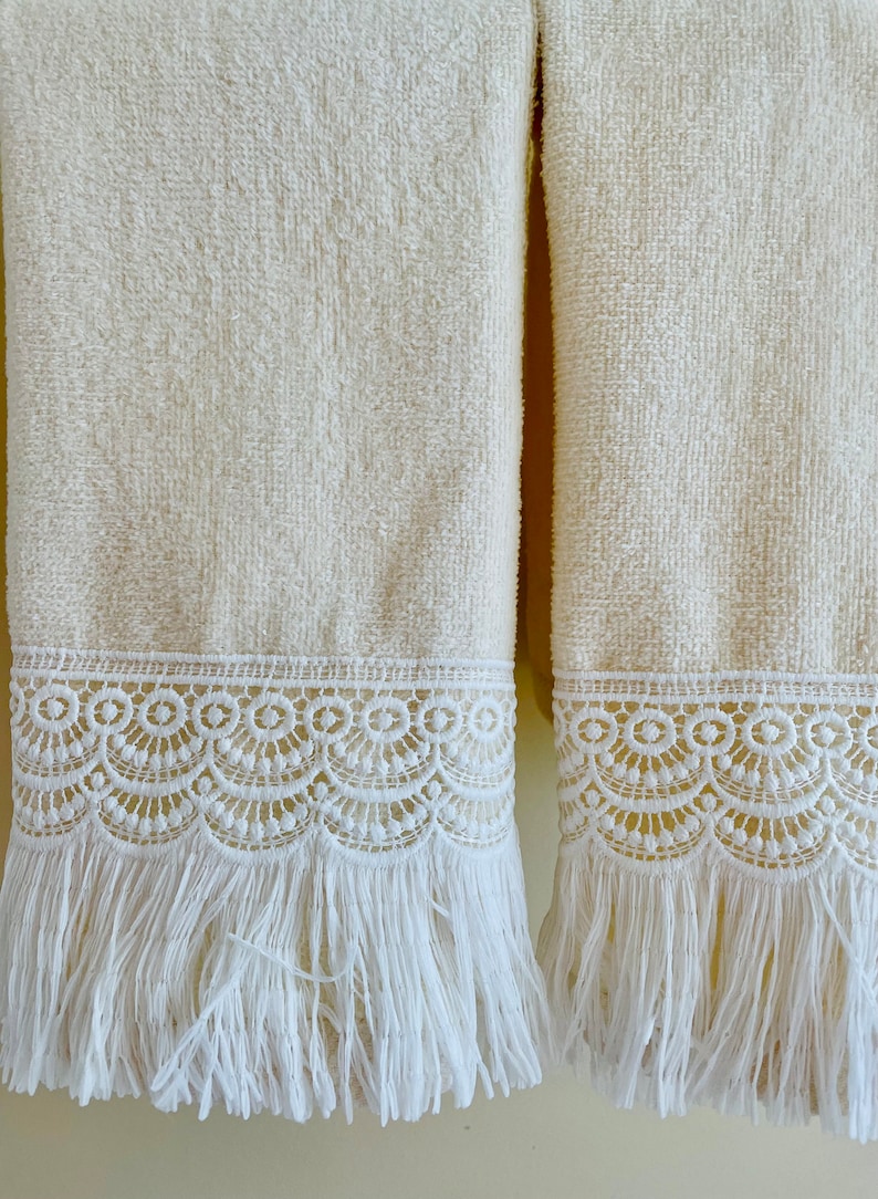 May include: Two cream-colored towels with white lace trim and fringe. The towels are hanging on a wall.