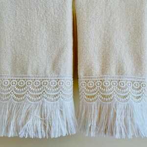 May include: Two white bath towels with white lace trim and fringe. The towels are hanging on a wall.