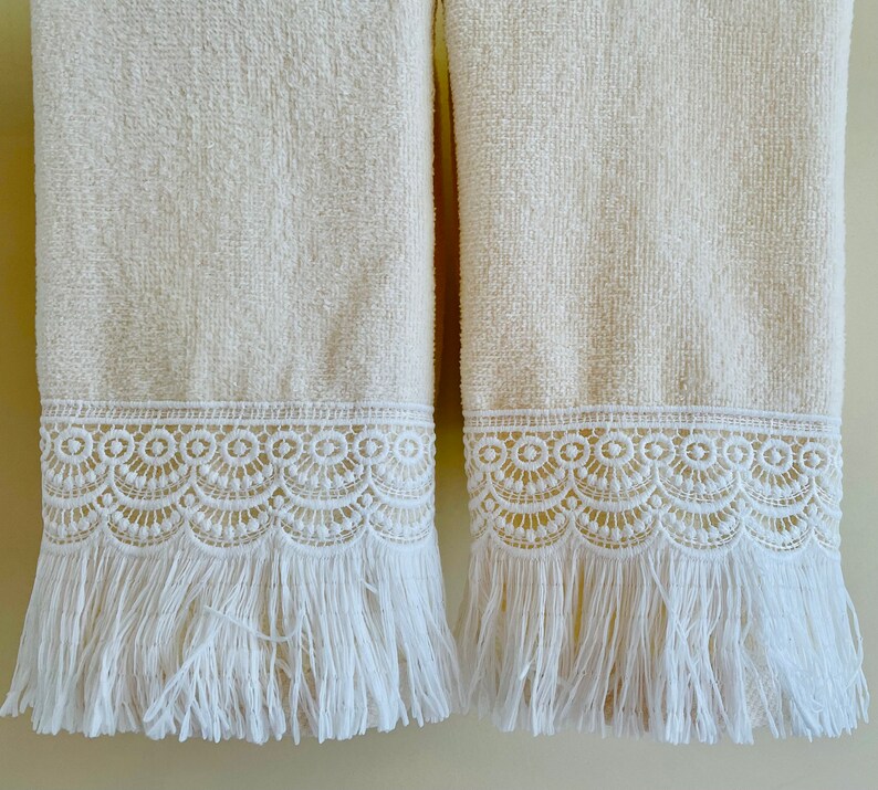 May include: Two white bath towels with white lace trim and fringe. The towels are hanging on a wall.