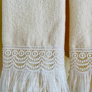 May include: Two white towels with white lace trim and fringe. The towels are hanging on a wall.