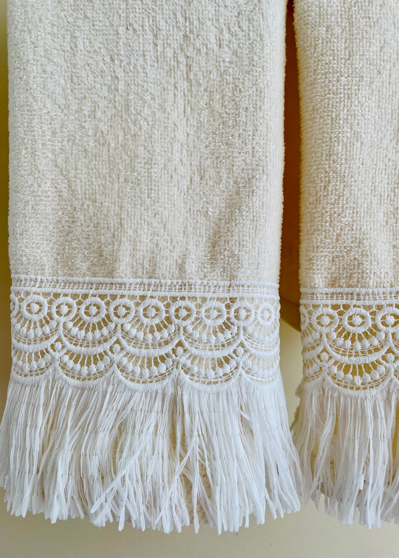May include: Two white towels with white lace trim and fringe. The towels are hanging on a wall.