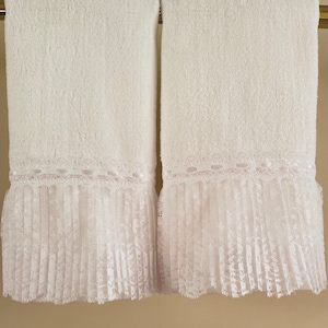 May include: Two white bath towels with a decorative lace and pleated trim. The towels are hanging on a gold-colored bar. The lace trim adds a touch of elegance to the otherwise simple design.