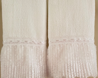 May include: Two white bath towels with a decorative lace and pleated trim. The towels are hanging on a gold-colored bar. The lace trim adds a touch of elegance to the otherwise simple design.