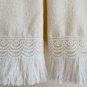 May include: Two cream-colored towels with white lace trim and fringe. The towels have a soft, fluffy texture.