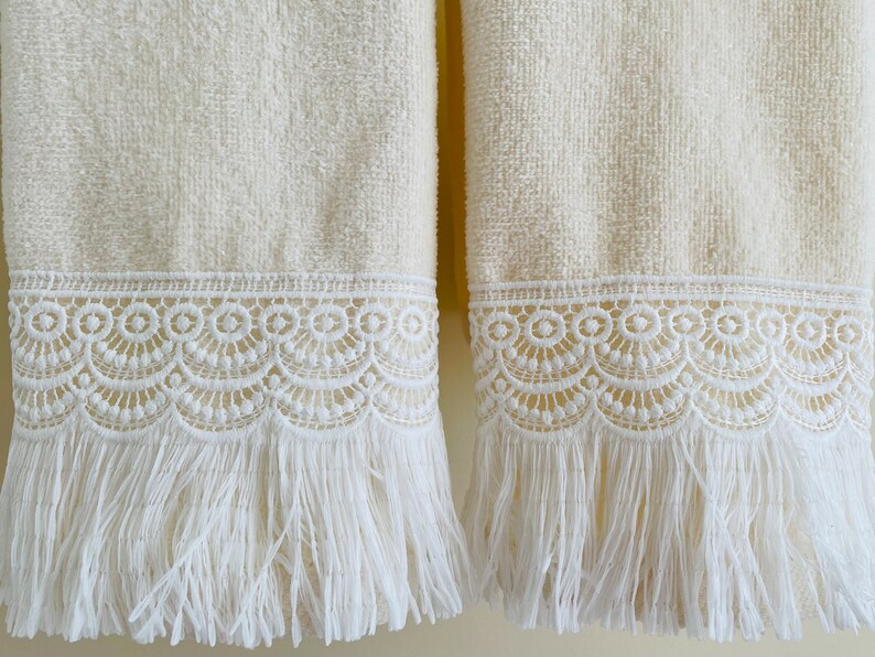 May include: Two cream-colored towels with white lace trim and fringe. The towels have a soft, fluffy texture.