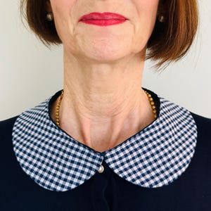 Op de afbeelding: Een marineblauwe cardigan met een afneembare Peter Pan-kraag. De kraag heeft een klassiek blauw-wit ruitpatroon. Een gouden ketting is zichtbaar. Een parelknop bevestigt de kraag aan de cardigan.