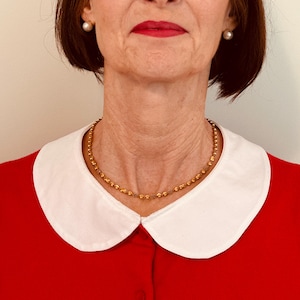 May include: A woman wearing a red collared shirt with a white collar and a gold chain necklace. She is wearing pearl earrings.