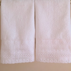 May include: Two white bath towels with white embroidered lace trim. The towels are hanging on a gold bar.