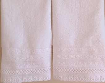 May include: Two white bath towels with white embroidered lace trim. The towels are hanging on a gold bar.