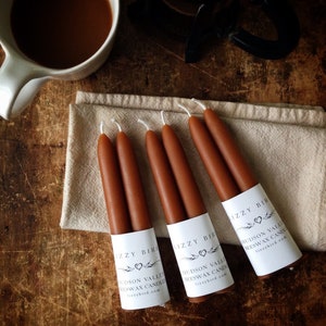 May include: Four pairs of brown beeswax candles with white labels that say "Izzy Bird" and "Hudson Valley Beeswax Candles" with a website address.