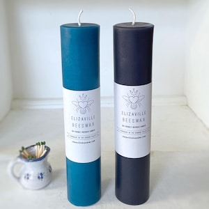 May include: Two cylindrical beeswax candles, one teal and one navy, with white labels featuring a bee and heart logo. The candles have a wick at the top and are labeled "Elizaville Beeswax". A small ceramic pitcher with matches is in the foreground.