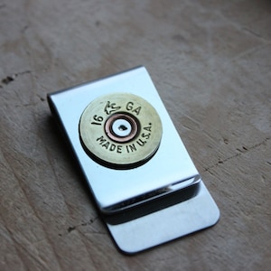 Może przedstawiać: Srebrny klips do pieniędzy z mosiężną łuską po naboju do strzelby. Łuska jest ostemplowana napisem "16 GA" i "MADE IN U.S.A.". Klips jest prostokątny z zaokrąglonymi rogami, na drewnianej powierzchni.