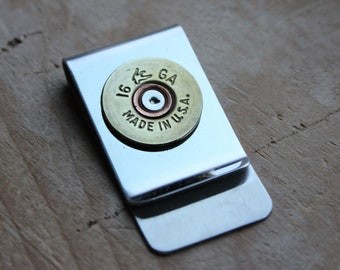 May include: A silver-colored money clip with a brass-colored shotgun shell casing attached. The casing is stamped with "16 GA" and "MADE IN U.S.A.". The clip is rectangular with rounded corners and is set against a wooden surface.