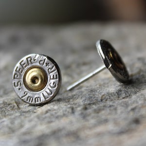 May include: A pair of silver-toned stud earrings with a bullet casing design. Each earring features a gold-colored center and the text "SPEER + LUGER 9mm". The earrings are displayed on a gray, textured surface.