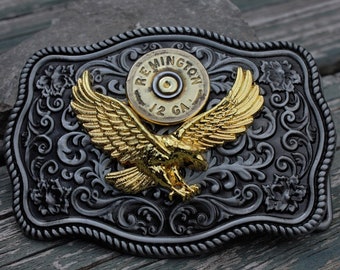 Peut inclure: Une boucle de ceinture en argent et or avec un motif d'aigle et une cartouche de fusil de chasse Remington 12 GA au centre.