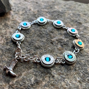 Puede incluir: Pulsera plateada con casquillos de bala con detalles turquesa. La pulsera incluye un pequeño dije de bota de vaquero. Los casquillos de bala están marcados con el texto "40 S&W WINCHESTER".