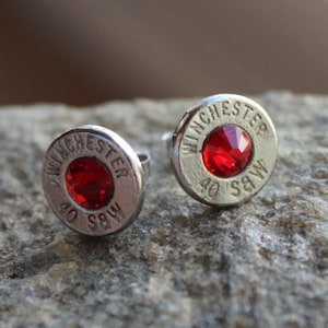 May include: A pair of silver bullet earrings with red crystal accents. The earrings are engraved with "Winchester 40 S&W".