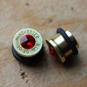 May include: Two brass-colored ear plugs with a red jewel center. One plug has the text "Winchester 30-30 WIN" engraved on it. The plugs have a black rubber ring around the edge.