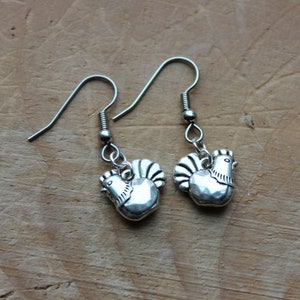May include: A pair of silver earrings featuring a small, detailed rooster charm. The earrings are made of metal and have a simple hook design.
