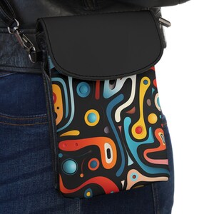 Puede incluir: Una bolsa de teléfono de cuero negro con correa cruzada con un patrón abstracto colorido. El patrón presenta remolinos, líneas y círculos en tonos de azul, rojo, amarillo y naranja sobre un fondo negro.