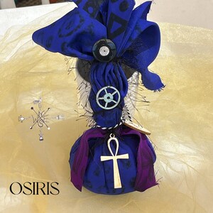 May include: A decorative object with a blue fabric bow featuring black shapes, a black button, and a silver charm. The object has a purple and blue fabric base with a gold ankh pendant. The word "OSIRIS" is at the bottom.