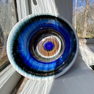 Puede incluir: Un cuenco de cerámica vidriada con un diseño de círculos concéntricos. El borde exterior es blanco, seguido de azul oscuro, azul claro y un centro marrón. El cuenco está colocado sobre un alféizar blanco, con un fondo natural.