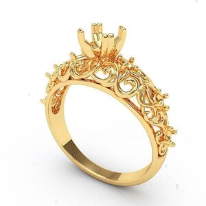 May include: A gold-colored ring with an ornate, filigree design. The ring features a detailed, openwork pattern along the band and a prong setting at the top, ready to hold a gemstone. The ring is set against a white background.