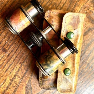 May include: Vintage opera glasses with a brown and gold finish, resting on a wooden surface. The binoculars are paired with a worn, tan leather case with green button closures. The lenses are black and silver.