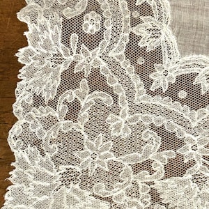 May include: Detailed close-up of a white lace textile with elaborate floral and leaf motifs. The lace has an openwork design, revealing a fine mesh. The edges are scalloped, enhancing the fabric's delicate appearance. The textile is likely used for decorative purposes.