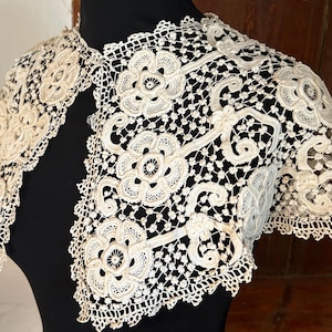 Könnte beinhalten: Elfenbeinfarbenes Spitzenbolero mit Blumen- und Rankenmustern. Das Bolero hat einen gewellten Rand und wird auf einer schwarzen Schaufensterpuppe präsentiert. Das aufwendige Design zeigt die filigrane Handwerkskunst des Kleidungsstücks.