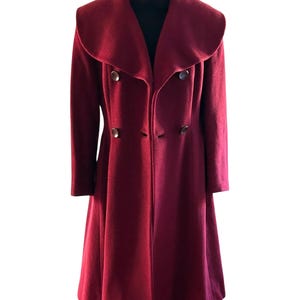 Puede incluir: Un abrigo largo burdeos con un cuello ancho y redondeado y botones grandes. El abrigo es de doble botonadura y tiene una falda acampanada. El abrigo se exhibe en un maniquí negro.