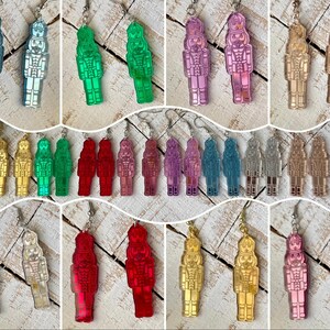 May include: A collection of colorful acrylic nutcracker earrings in various colors, including blue, green, pink, gold, and red. Each earring features a detailed nutcracker design with a silver-tone hook.