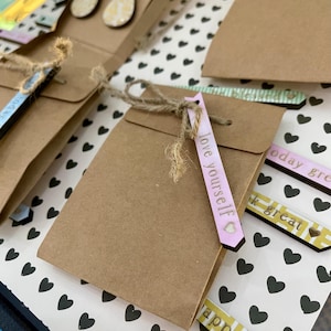 May include: Close-up of several small brown paper gift bags tied with twine. Each bag has a decorative tag with positive affirmations like "love yourself" and "happy". The background is a white surface with black heart patterns.
