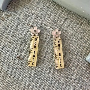 Puede incluir: Un par de pendientes colgantes. Cada pendiente presenta una flor rosa claro con un centro de pedrería. Debajo de la flor hay un dije en forma de regla con las palabras "YOU RULE" impresas verticalmente en negro.