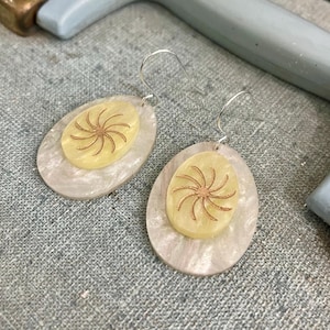 May include: A pair of dangle earrings. Each earring features a light yellow oval shape with a brown sunburst design, set on a larger, pearlescent oval. The earrings have silver-colored hooks.