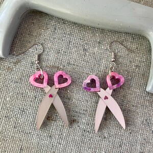 May include: A pair of silver-coloured earrings shaped like scissors, with pink heart-shaped handles. The earrings have a small heart detail where the blades cross and are suspended from silver hooks.