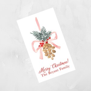 Puede incluir: Una ilustración de acuarela de una piña con un lazo rosa y bayas rojas. El texto "Merry Christmas! The Bryant Family" está escrito en rojo debajo de la piña.