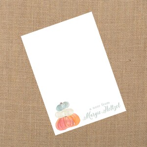 May include: A stack of three pumpkins in shades of orange, white, and blue. The pumpkins are stacked on top of each other and have a stem on top. The text "a note from Margie Heltzel" is written below the pumpkins.