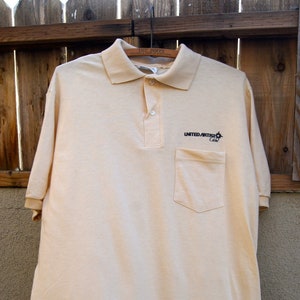 Könnte beinhalten: Ein hellbeiges Poloshirt mit einer Tasche auf der linken Seite. Das Shirt hat ein schwarzes Logo mit der Aufschrift "United Artists Cable".