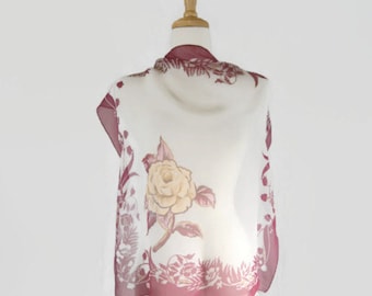 Puede incluir: Una bufanda de seda blanca con un diseño floral rosa con una rosa grande. La bufanda se muestra en un maniquí de madera.