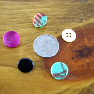 Op de afbeelding: Assortiment van kleurrijke knopen, waaronder een roze knoop, een zwarte knoop, een turquoise knoop, een gele knoop met drie gaten en een gepatenteerde knoop in de kleuren rood, wit en blauw. Een zilveren Amerikaanse dime-munt is ook inbegrepen.