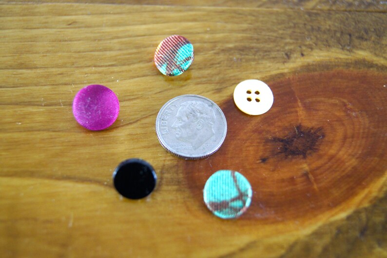 Op de afbeelding: Assortiment van kleurrijke knopen, waaronder een roze knoop, een zwarte knoop, een turquoise knoop, een gele knoop met drie gaten en een gepatenteerde knoop in de kleuren rood, wit en blauw. Een zilveren Amerikaanse dime-munt is ook inbegrepen.