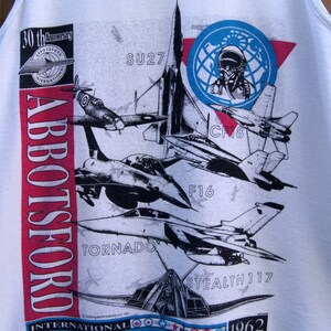 May include: White tank top with a red, white, and blue graphic of various fighter jets. The graphic includes the text "Abbotsford International Airshow 1962-1991".