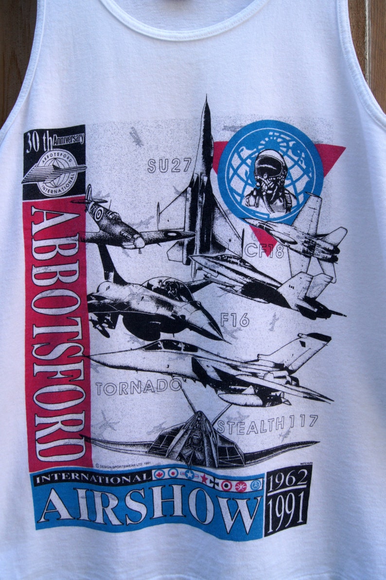 May include: White tank top with a red, white, and blue graphic of various fighter jets. The graphic includes the text "Abbotsford International Airshow 1962-1991".